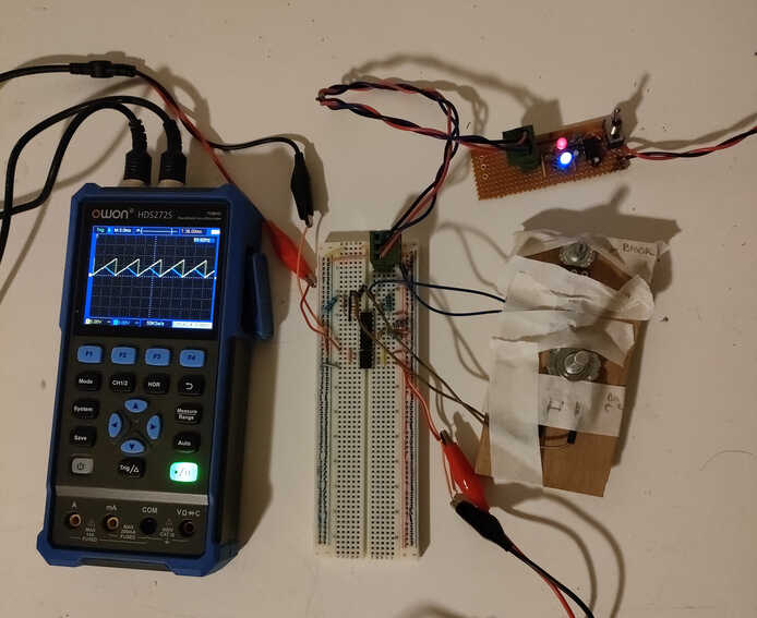 An oscilloscope connected to a breadboard circuit. The screen of the oscilloscope shows a triangle wave and sawtooth wave. Some knobs are taped to a sheet of cardboard and wired into the breadboard. An additional circuit with a red and blue LED is attached to the breadboard with a cable.