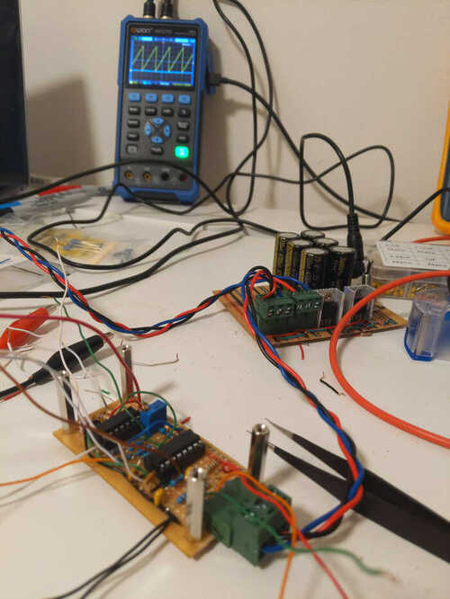 A pair of circuit boards connected by a DIY power cable with an oscillator in the background displaying a sawtooth wave